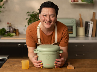 A person in an orange shirt and apron smiles while holding a green cooking pot on a wooden kitchen counter, with utensils and ingredients in the background.