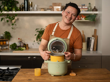 A man in an orange shirt and beige apron stands in a kitchen, smiling while placing a yellow container into a green rice cooker on the counter.