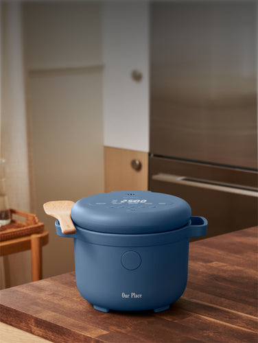 A blue electric cooker with a wooden handle sits on a kitchen countertop next to a potted plant and a bottle of oil.