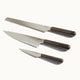 Three kitchen knives with dark handles are arranged in a row on a white background: a bread knife, a chef’s knife, and a paring knife.