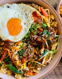 A bowl of bibimbap topped with a sunny-side-up egg, sliced beef, vegetables, rice, gochujang sauce, and sprinkled sesame seeds.