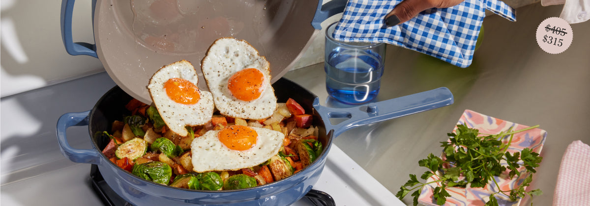Translation missing: en.A person adds three sunny-side-up eggs from a pan onto a skillet of roasted vegetables on a stove, holding the pan with a blue checkered oven mitt