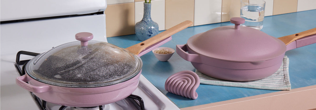 Translation missing: en.Two pink nonstick pans with wooden handles are on a stove and countertop, one with a glass lid. Nearby are a striped towel, a silicone trivet, a bowl, a vase, and a glass of water