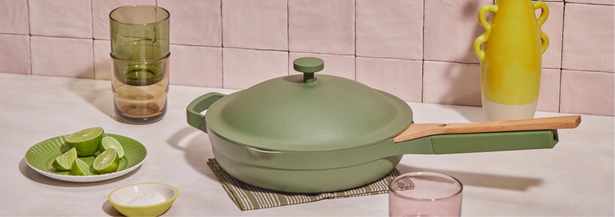 Translation missing: en.A green multipurpose pan with a wooden handle sits on a mat; nearby are stacked glasses, a plate with lime wedges, a yellow vase, and a small bowl of salt