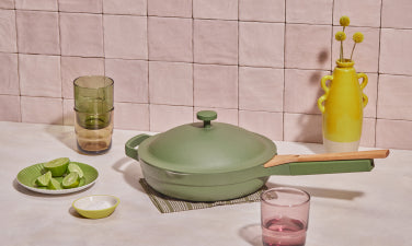Translation missing: en.A green pan with a wooden handle sits on a table next to drinking glasses, a plate of lime wedges, a small bowl, and a yellow vase with flowers against a tiled background