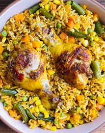 A bowl of seasoned rice mixed with peas, corn, carrots, and green beans, topped with two cooked chicken drumsticks.