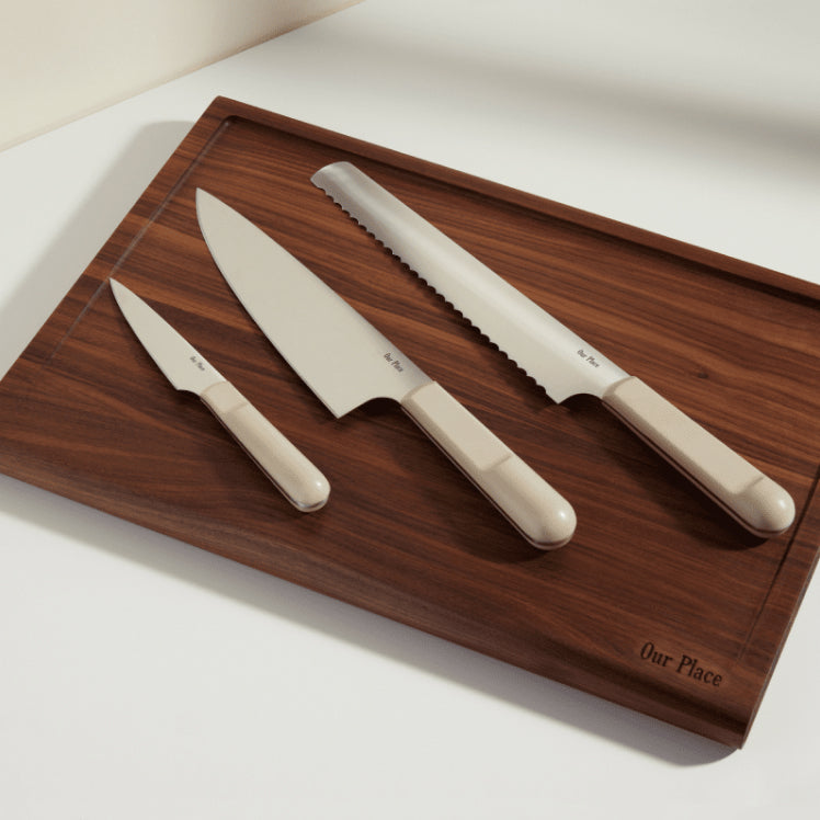 beige chefs knife, serrated knife, and paring knife on walnut cutting board