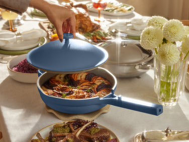 A hand lifts the lid of a blue pan, revealing a baked dish with sliced vegetables on a set dining table with various dishes and white flowers.