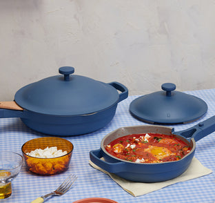 A blue pan with a tomato-based dish sits on a blue checkered tablecloth beside its lid, another pan, a drink, a bowl of cheese, and a fork on a napkin.