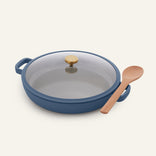 The Dual Handle Always Pan in blue, featuring a shallow nonstick design with a glass lid and gold knob, is pictured beside a wooden spoon on an off-white background. This versatile and non-toxic pan is ideal for everyday cooking.