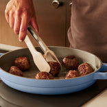 A person uses tongs to cook several meatballs in a light blue skillet on a stovetop.