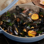 Mussels steaming in a blue pan with fresh herbs and halved grilled lemons.