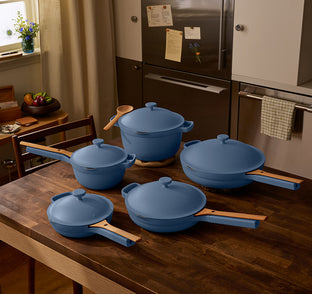 Four blue cookware pots and pans with wooden handles are arranged on a wooden kitchen table in a modern kitchen.