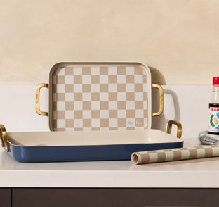 A checkered baking dish set with gold handles is displayed on a countertop, accompanied by matching patterned paper and a bottle of sauce.