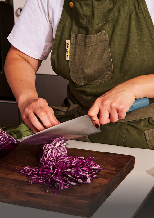 A person wearing a green apron slices red cabbage on a wooden cutting board with a large knife.