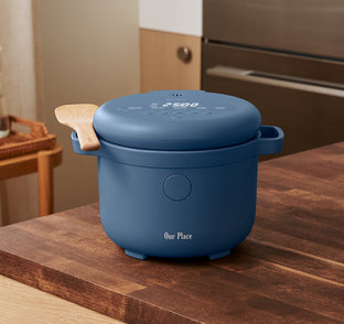 A blue electric rice cooker labeled Our Place sits on a wooden kitchen counter, with a wooden spatula resting on its side handle.