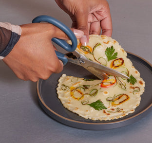 A person uses kitchen scissors to cut a flatbread topped with herbs and sliced peppers on a dark plate.