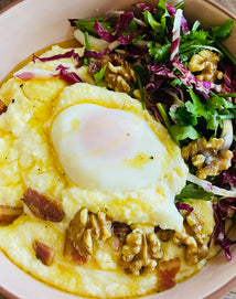 A bowl of creamy polenta topped with a poached egg, walnuts, bacon pieces, and served with mixed greens.