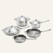 A five-piece stainless steel cookware set with glass lids and long handles, arranged on a light beige background.