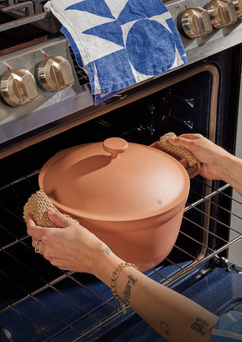 A terracotta clay Dutch oven with a lid is being placed into a hot oven using textured brown oven mitts. A blue and white patterned kitchen towel is draped over the stove handle in the background.