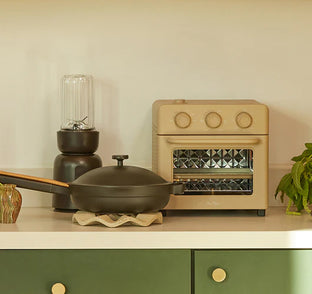 A beige toaster oven, black blender, and black pan with a wooden handle are arranged on a white countertop with green cabinetry below.