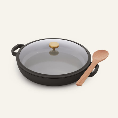 Versatile and non-toxic, the black Dual Handle Always Pan—with a glass lid and an elegant gold knob—is pictured alongside a wooden spoon on a plain light background.