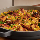 Close-up of a pan filled with chicken biryani, showing pieces of chicken, yellow rice, peas, nuts, and whole spices like cinnamon sticks and star anise.
