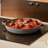 A shallow gray pan filled with spaghetti, tomato sauce, and meatballs sits on a stovetop in a modern kitchen.