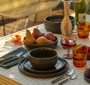 A table set for a meal with black dishes, pears in a bowl, glasses with drinks, forks, spoons, napkins, and a bottle on a striped tablecloth.