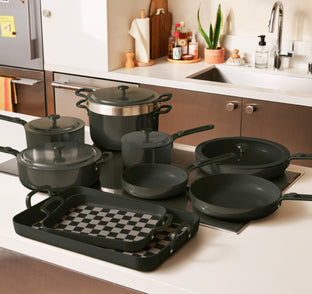 A set of black pots, pans, and a checkered baking tray are arranged on a kitchen counter near a sink and stove.