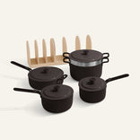 The Essentials Saucepan Set includes four black pots and pans with lids, a stainless steel steamer insert, and a beige vertical organizer rack with nesting storage, all displayed on a light background.