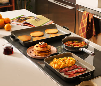 A kitchen counter with pancakes, scrambled eggs, bacon, and sautéed vegetables on stovetop pans, a stack of pancakes on a plate, oranges, an open cookbook, and syrup nearby.