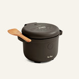 An Our Place Rice Cooker in matte black with a digital display, topped with a wooden spatula, set against a plain light background.