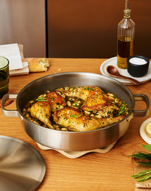 Roasted chicken pieces with herbs and white beans in a stainless steel pan, placed on a wooden table with olive oil, salt, bread, a green glass, and a lemon slice nearby.