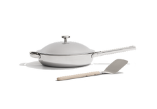 A stainless steel frying pan with a lid and a long handle is placed next to a metal spatula with a light-colored handle on a plain white background.