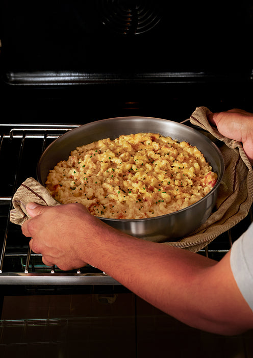 Person using oven mitts to place a round pan of baked macaroni and cheese with a breadcrumb topping onto an oven rack.