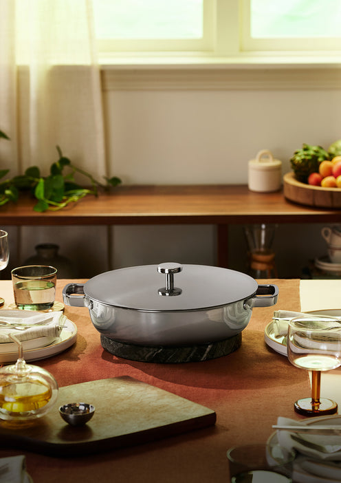 A lidded stainless steel pot sits on a trivet at the center of a set dining table with glassware, plates, and food, in a sunlit room.