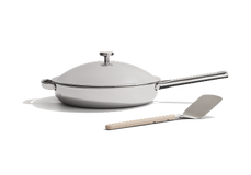 A stainless steel frying pan with a lid and a long handle is shown next to a metal spatula with a light-colored handle against a white background.