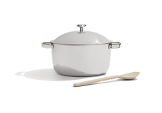 A stainless steel pot with handles and a lid is placed next to a beige wooden spoon on a white background.