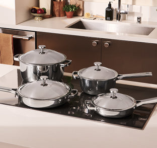 Four stainless steel pots and pans with lids are arranged on a modern black glass stovetop in a clean kitchen setting.