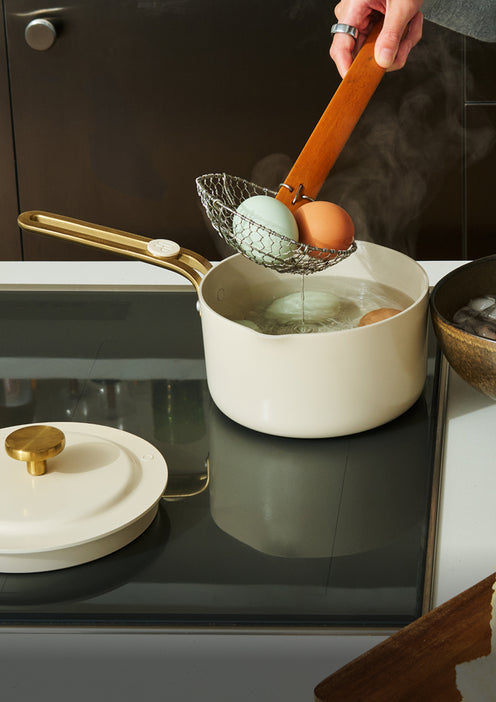 A hand uses a wire skimmer to remove three eggs—brown, blue, and light brown—from a pot of boiling water on a stovetop.