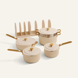 The Essentials Saucepan Set includes three cream-colored, lidded nonstick saucepans with gold handles, a matching lid rack, and a nesting storage system for easy organization.