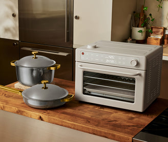 A countertop oven with a digital display sits on a wooden counter next to two gray pots with gold handles and lids in a modern kitchen.