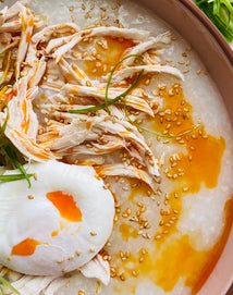 A bowl of congee topped with shredded chicken, a poached egg, sesame seeds, sliced scallions, and a drizzle of chili oil.