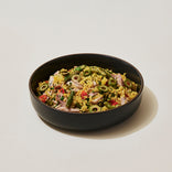 A black bowl filled with rice mixed with shredded meat, green beans, olives, tomatoes, and herbs, set on a light surface.