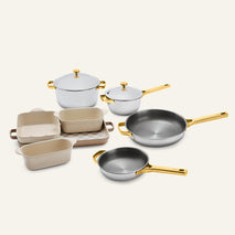 The Titanium Pro Cookware + Bakeware Set (2.0) featuring metal pots and pans with gold-toned handles and lids, and three light-colored baking dishes on a tray.