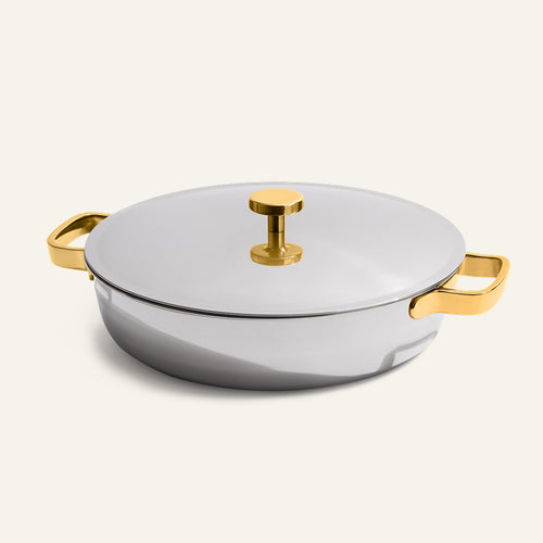 A round silver Titanium Braiser Pro 28 cm stainless steel casserole dish with a lid, featuring chef-approved gold-colored handles and a matching gold knob on top, is placed on a plain off-white background.