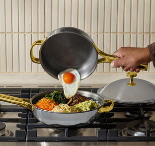 A hand pours a fried egg from a small pan onto a skillet filled with vegetables and meat on a stovetop.