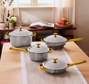 A set of stainless steel cookware with gold handles and lids, displayed on a wooden table in a bright kitchen setting.