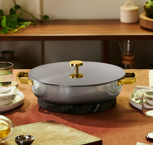 A gray and gold pot with a lid sits on a dark trivet, centered on a set dining table with plates, glasses, and napkins.